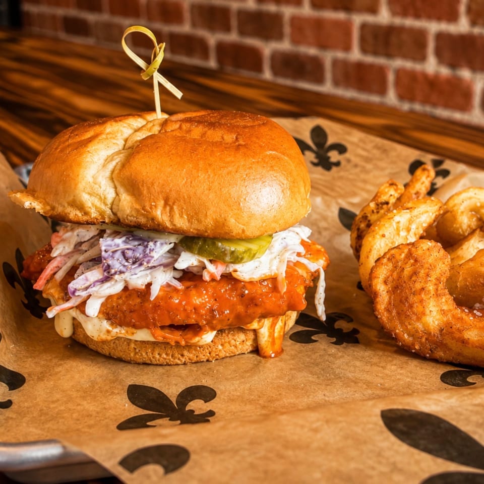 Spicy chicken sandwich with coleslaw and pickles, served with pretzel bites on branded paper.